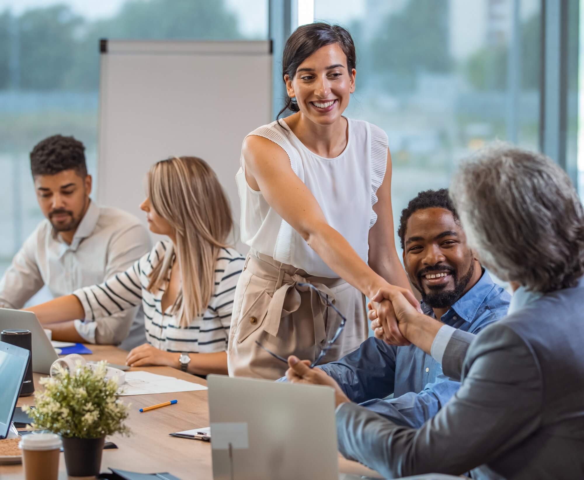 A woman, standing, shakes hands with a man, sitting at a table filled with people working.